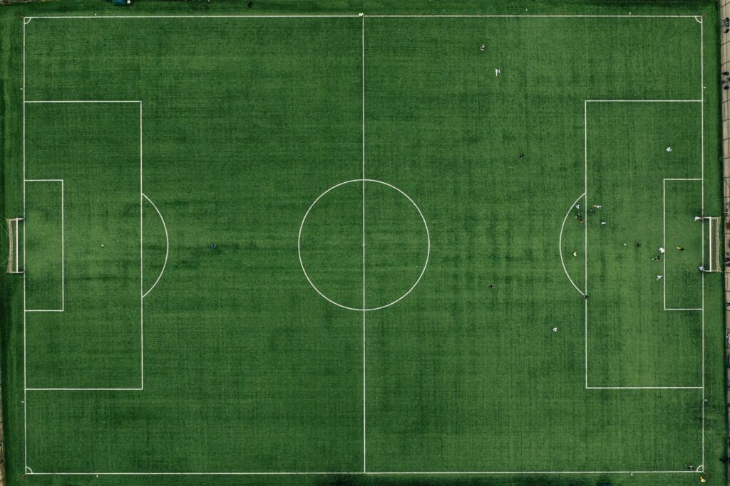 Aerial view of a green soccer field with white markings. A group of players is gathered near the right goal, while the rest of the field is mostly empty.