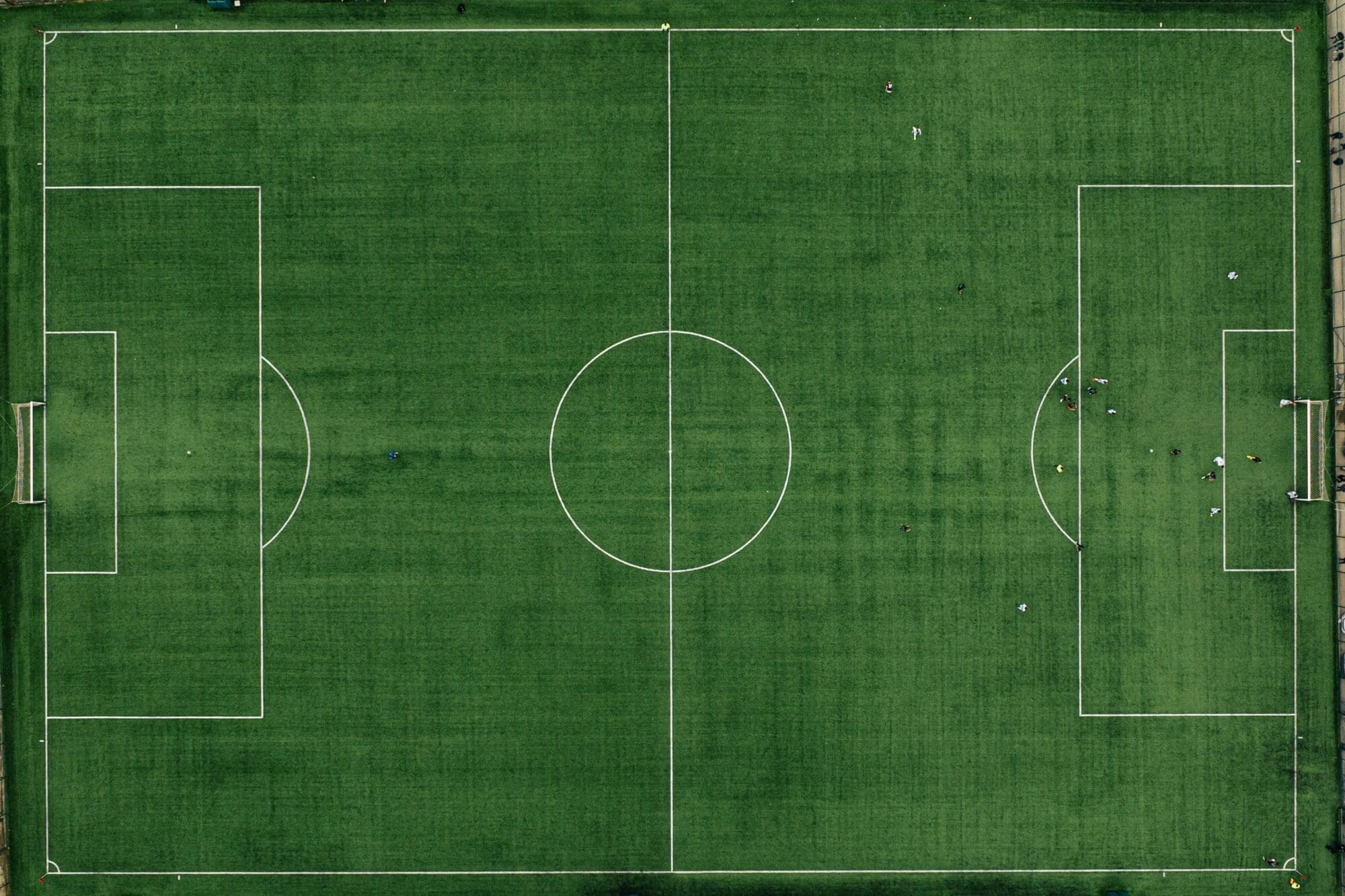 Aerial view of a green soccer field with white markings. A group of players is gathered near the right goal, while the rest of the field is mostly empty.