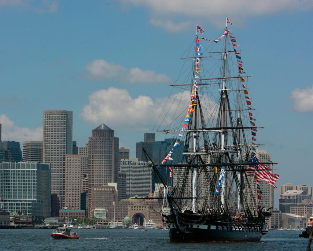 A historic tall ship decorated with colorful flags sails on water, with a small boat nearby and a city skyline with modern skyscrapers in the background under a partly cloudy sky.