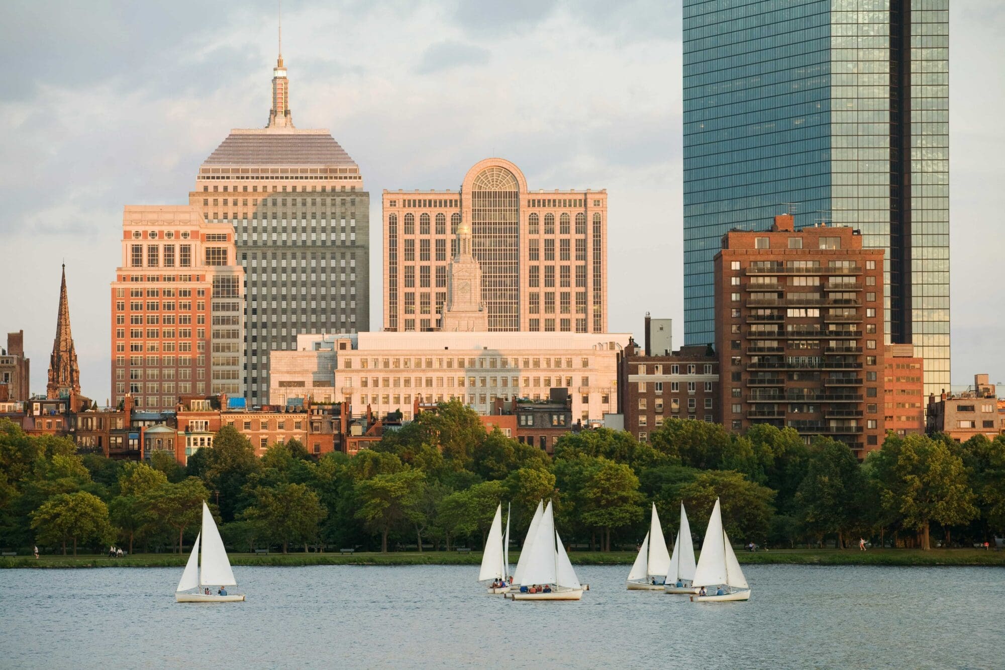 White sailboats float on a river with a tree-lined shore in the foreground and tall city buildings and skyscrapers in the background under a partly cloudy sky.