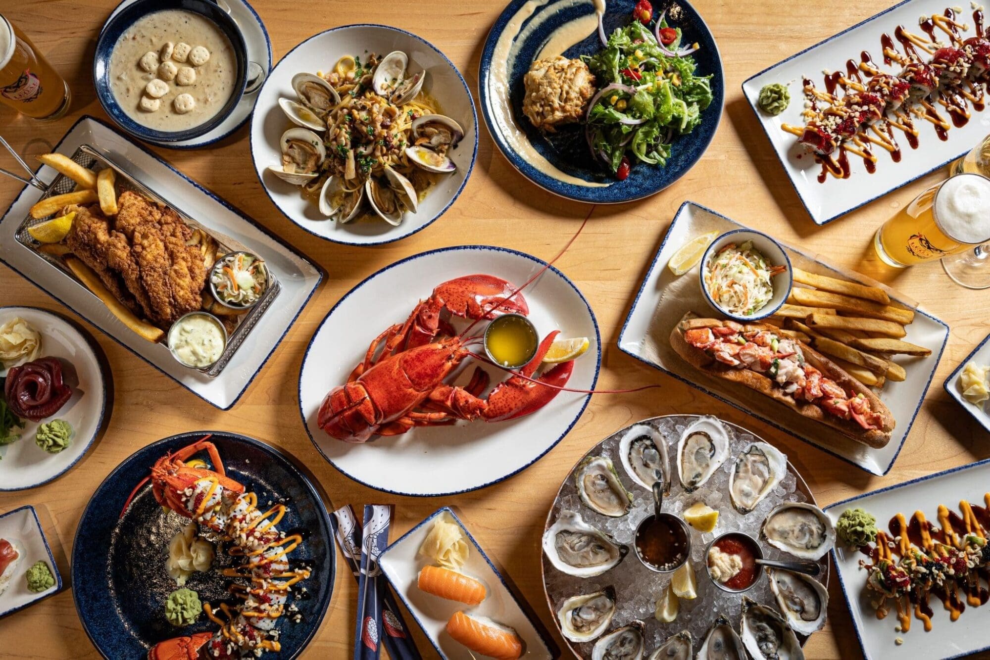 A table set with a variety of seafood dishes, including a whole lobster, oysters, clams, fried fish with fries, lobster rolls, sushi, soup, salad, and beer, all arranged on wooden surface.