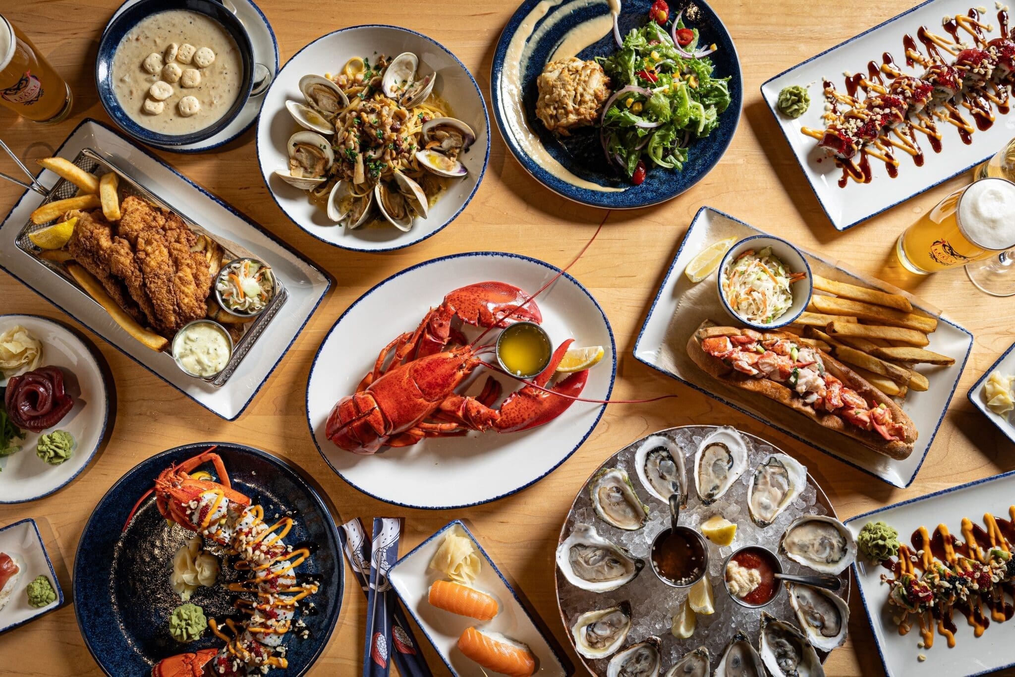 A table set with a variety of seafood dishes, including a whole lobster, oysters, clams, fried fish with fries, lobster rolls, sushi, soup, salad, and beer, all arranged on wooden surface.