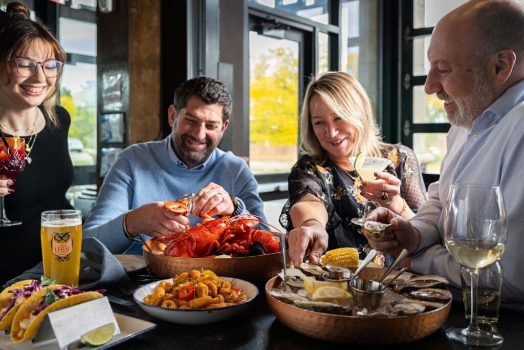 Four people happily share a seafood meal at a restaurant table, enjoying lobster, oysters, fried calamari, tacos, drinks, and corn, with light streaming in from large windows behind them.