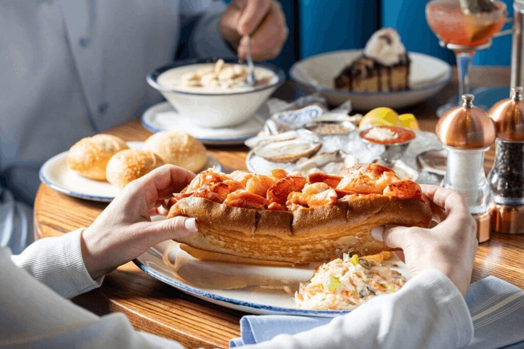 A person holds a lobster roll with coleslaw on the side; in the background are bread rolls, a bowl of soup, oysters on ice, and a slice of chocolate cake on a wooden table.