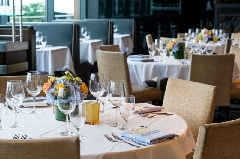 A restaurant dining area with round tables covered in white tablecloths, set with glassware, cutlery, and floral centerpieces. Beige chairs surround the tables, and natural light enters through large windows.