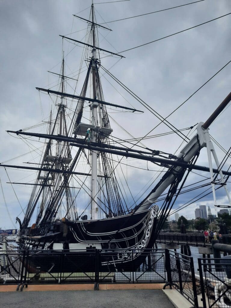 A large historic wooden sailing ship with tall masts and rigging is docked at a pier under a cloudy sky, with a city skyline visible in the background.