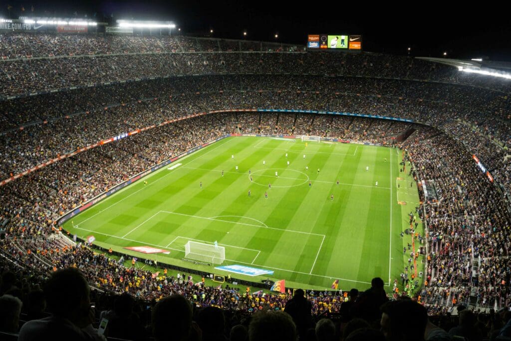 A packed soccer stadium at night with thousands of fans watching a match in progress on a brightly lit field. The teams are spread out across the pitch and the stands are filled with spectators.