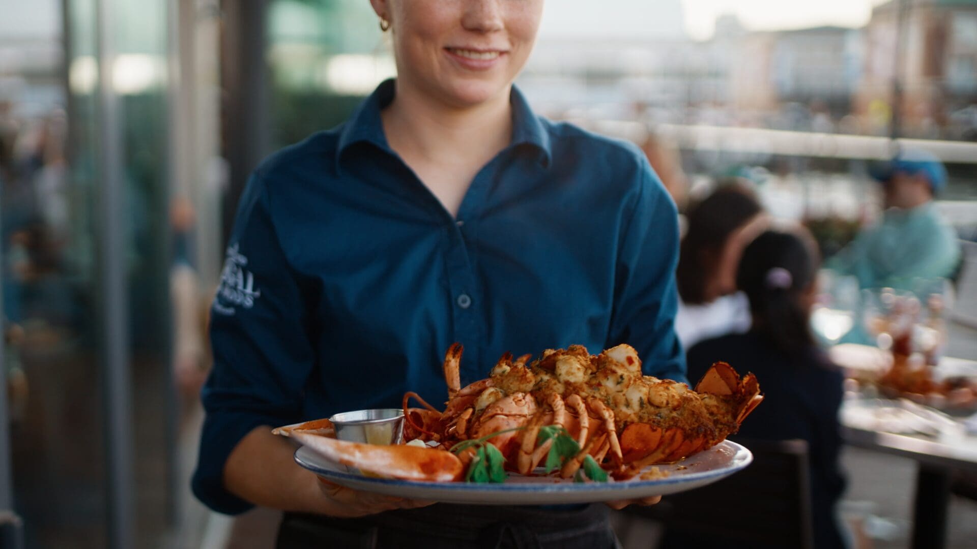 A server in a blue shirt holds a large plate with a cooked lobster, sauce, and garnish at an outdoor restaurant with people dining in the background.