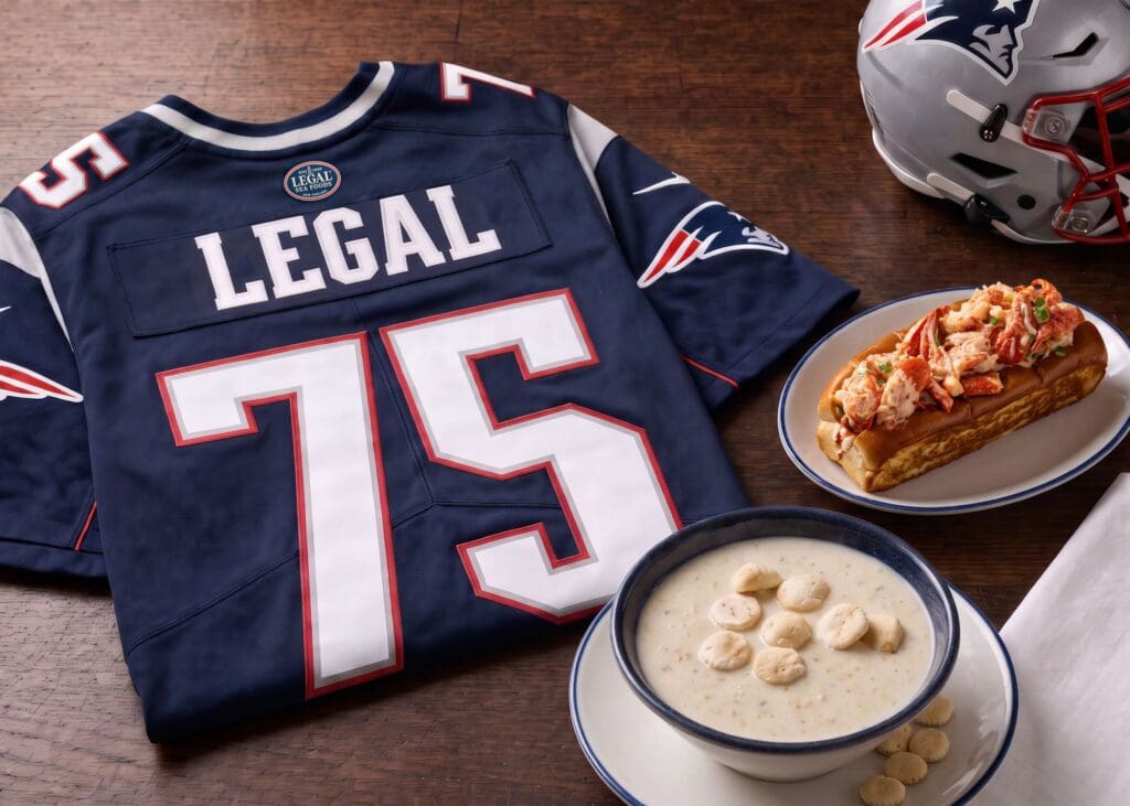 A New England Patriots jersey with “LEGAL 75” on it sits next to a lobster roll, a bowl of clam chowder with oyster crackers, and a football helmet on a wooden table.