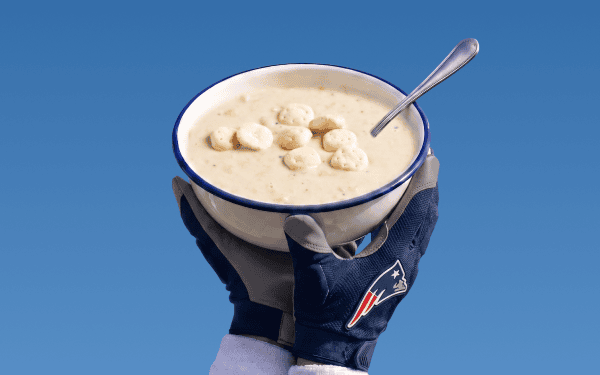 A person wearing New England Patriots gloves holds a bowl of clam chowder topped with oyster crackers and a spoon, against a blue background.