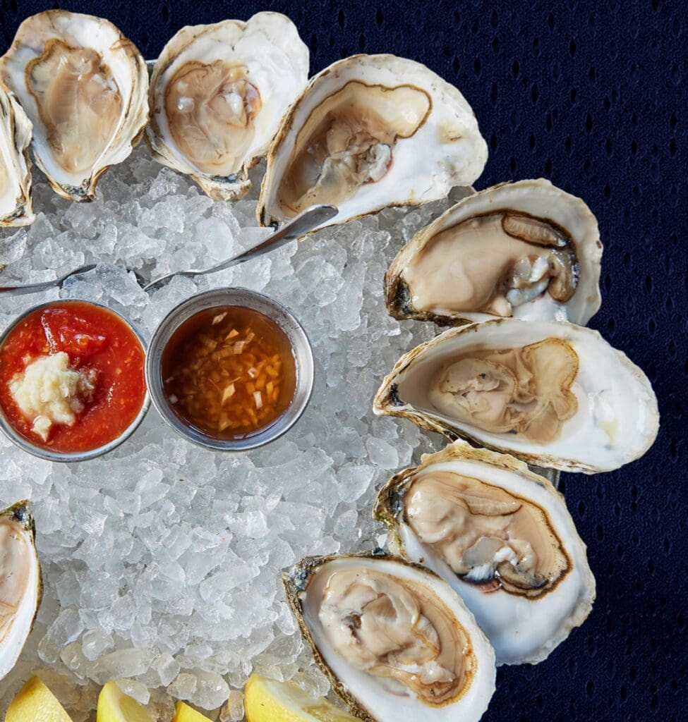 A platter of oysters on ice, served with cocktail sauce, minced onion sauce, lemon wedges, and small metal spoons, set against a dark blue cloth background—perfect for savoring at a Patriots watch party on a Boston rooftop.