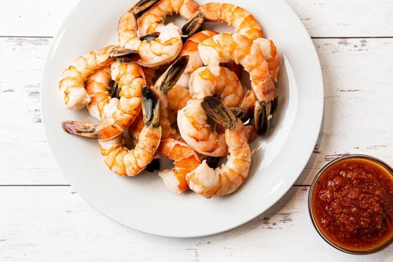 A white plate with cooked shrimp arranged in a circle, next to a small bowl of cocktail sauce on a white wooden surface.