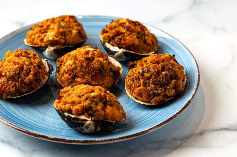 Five stuffed clams with a golden-brown breadcrumb and seafood filling are arranged on a blue plate with a white rim, set on a light marble surface.