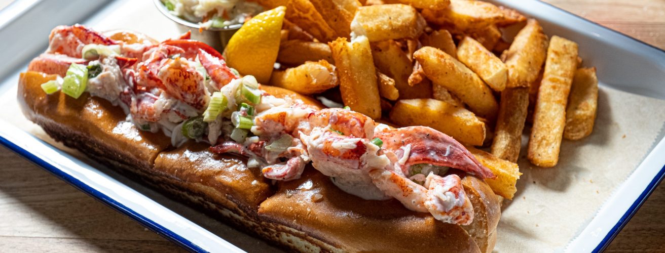 Half pound Maine lobster roll with lemon mayo, french fries and coleslaw on metal tray.